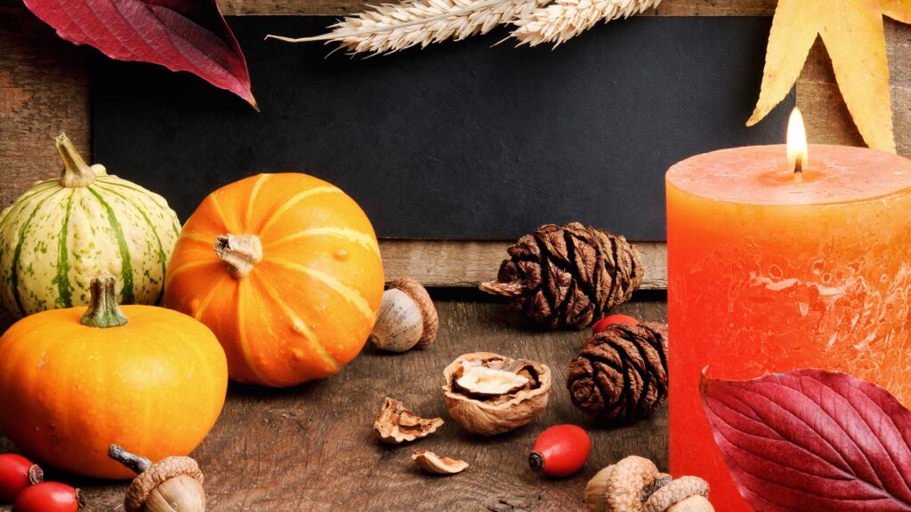 Das Bild zeigt eine herbstliche Szene mit verschiedenen Naturmaterialien, die als Schmuck dienen könnten. Zu sehen sind kleine Kürbisse in Orange und Grün, Tannenzapfen, Nüsse, Hagebutten und bunte Herbstblätter. Im Hintergrund befindet sich eine orangefarbene Kerze und einige Weizenähren. Die Elemente sind auf einem rustikalen Holztisch arrangiert.